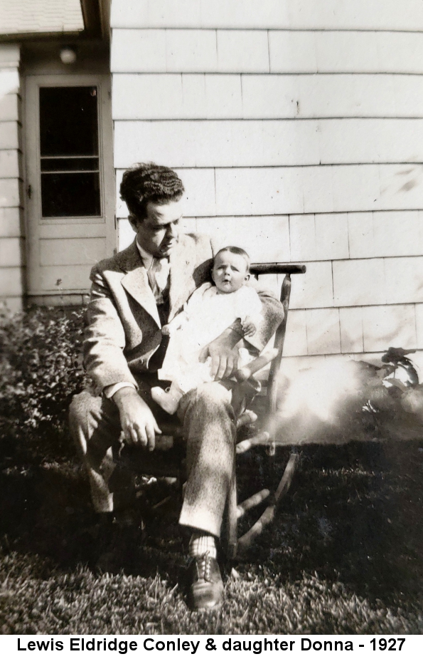 Black and white photo of a young man with thick curly hair, wearing a suit, and sitting in a rocking chair on the lawn outside a white shingled house, a baby in a dress on his lap.